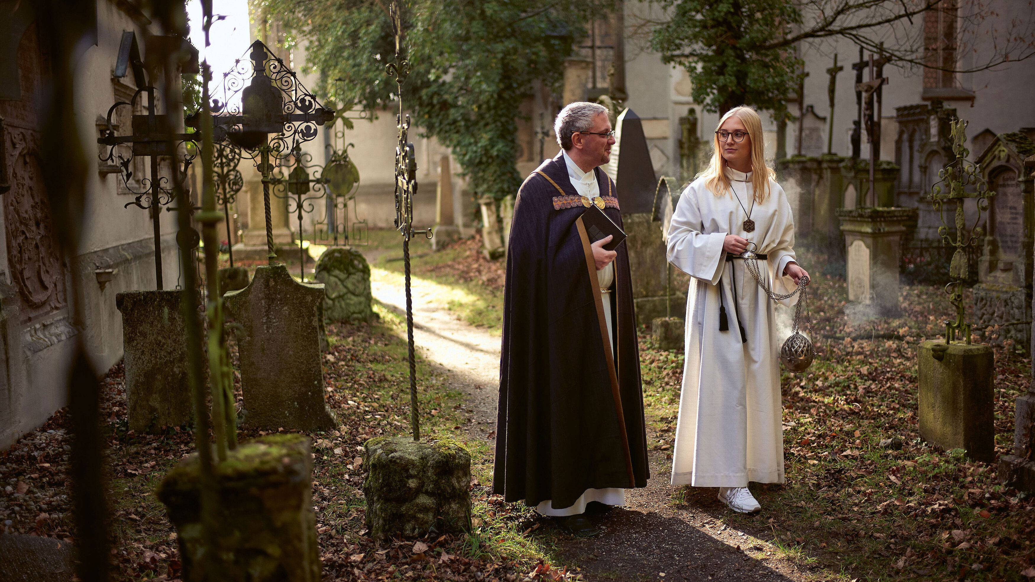 Ein Priester und eine Ministrantin mit Weihrauch gehen über einen Friedhof und unterhalten sich.