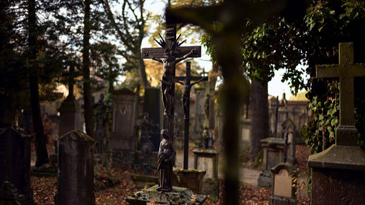 Ein Friedhof im Herbst. Blick über die Gräber hinweg auf eine Jesusfigur am Kreuz.