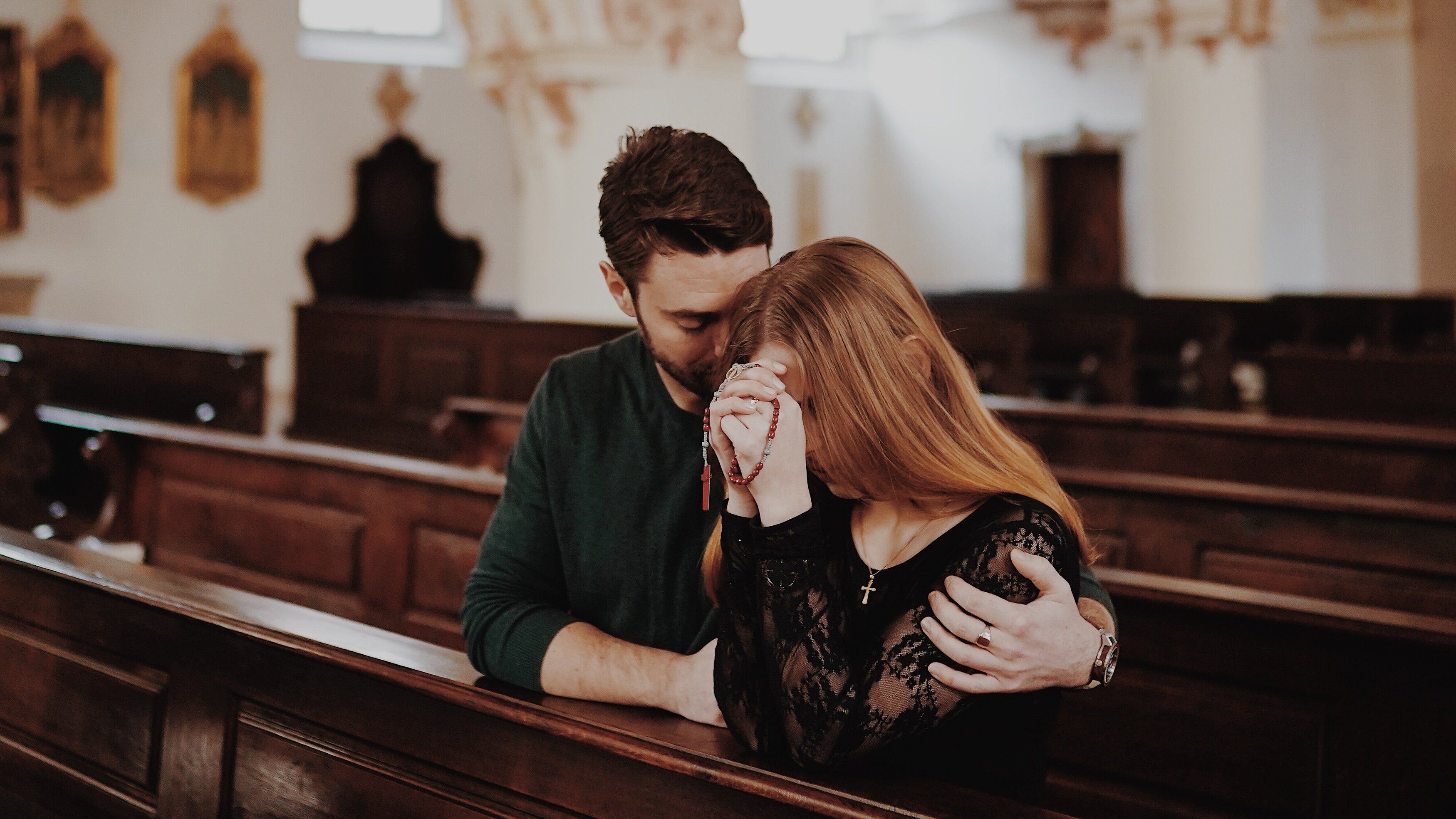 Ein junger Mann und eine junge Frau sitzen in einer Kirche. Er hat den Arm tröstend um sie gelegt, während sie den Rosenkranz betet.