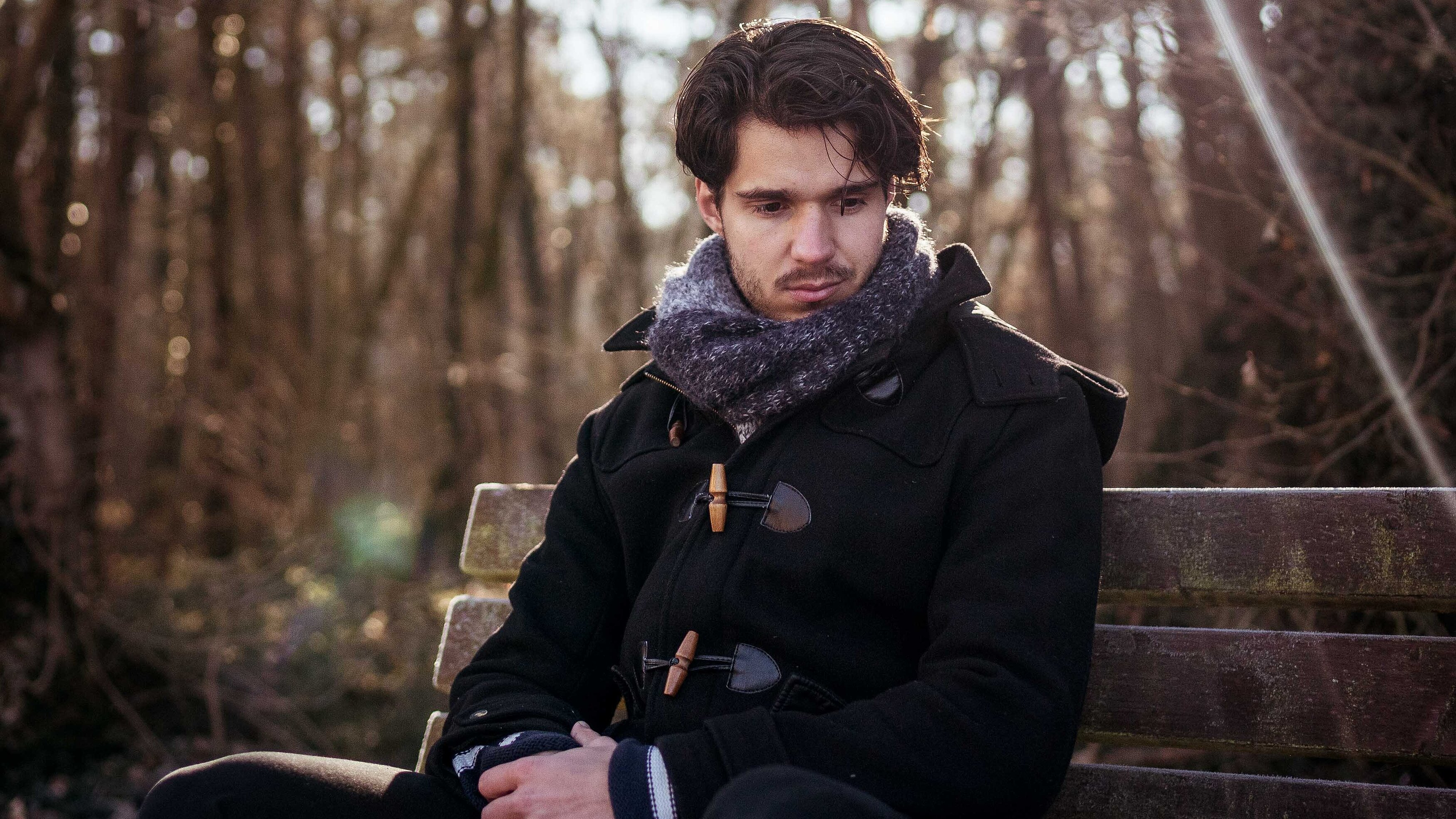 Ein junger Mann sitzt auf einer Bank im Wald und schaut zu Boden.