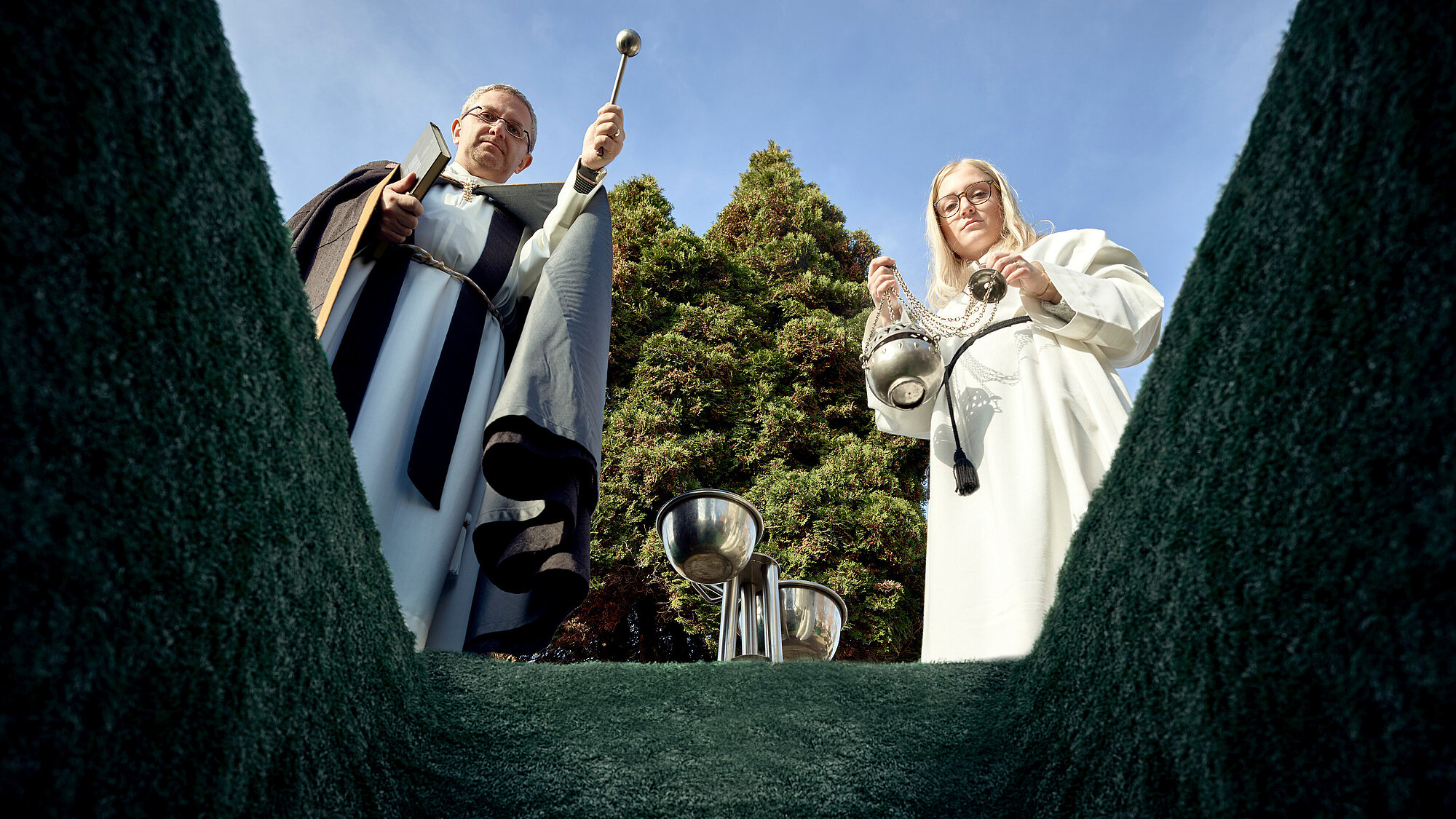 Blick aus einer Sarggrube nach oben auf einen Priester, der gerade die Segnung vornimmt und eine Ministrantin mit Weihrauch neben ihm.