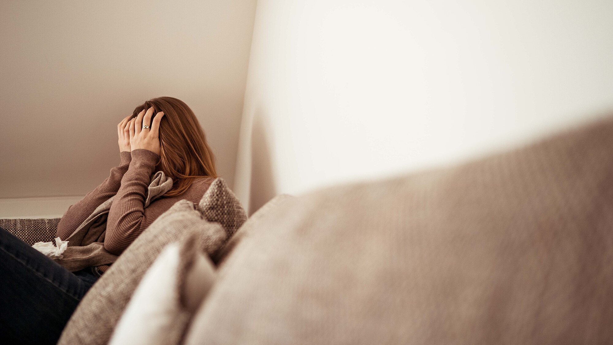 Eine Frau sitzt in der Ecke einer Couch und hat ihre Hände vor dem Gesicht verschlagen.