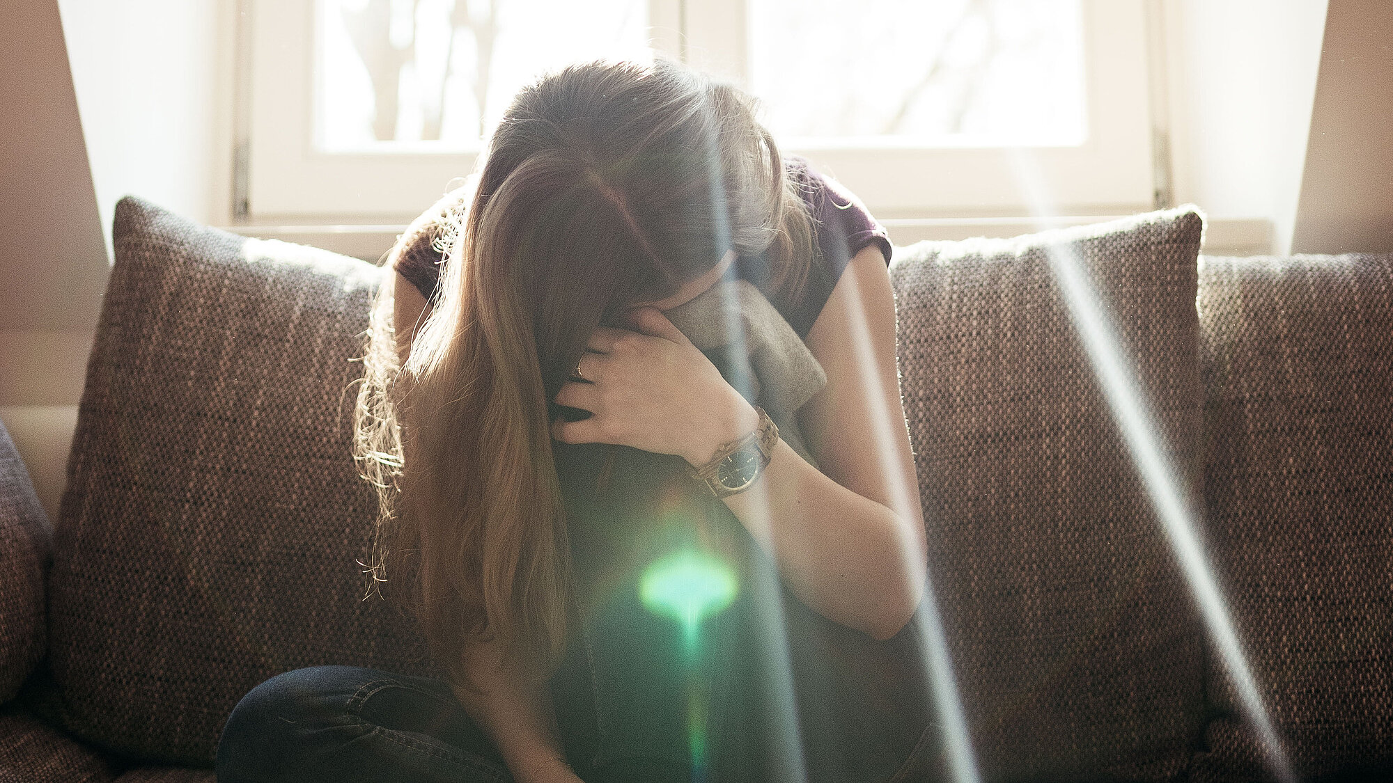 Eine junge Frau sitzt zusammengekauert vor einem Fenster und hat das Gesicht in einem Pulli ihres verstorbenen Freundes begraben.