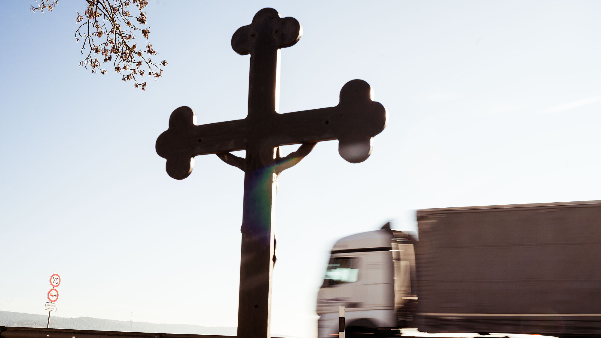 Ein Kreuz am Straßenrand im Fokus, im Hintergrund fährt ein Lastwagen vorbei.